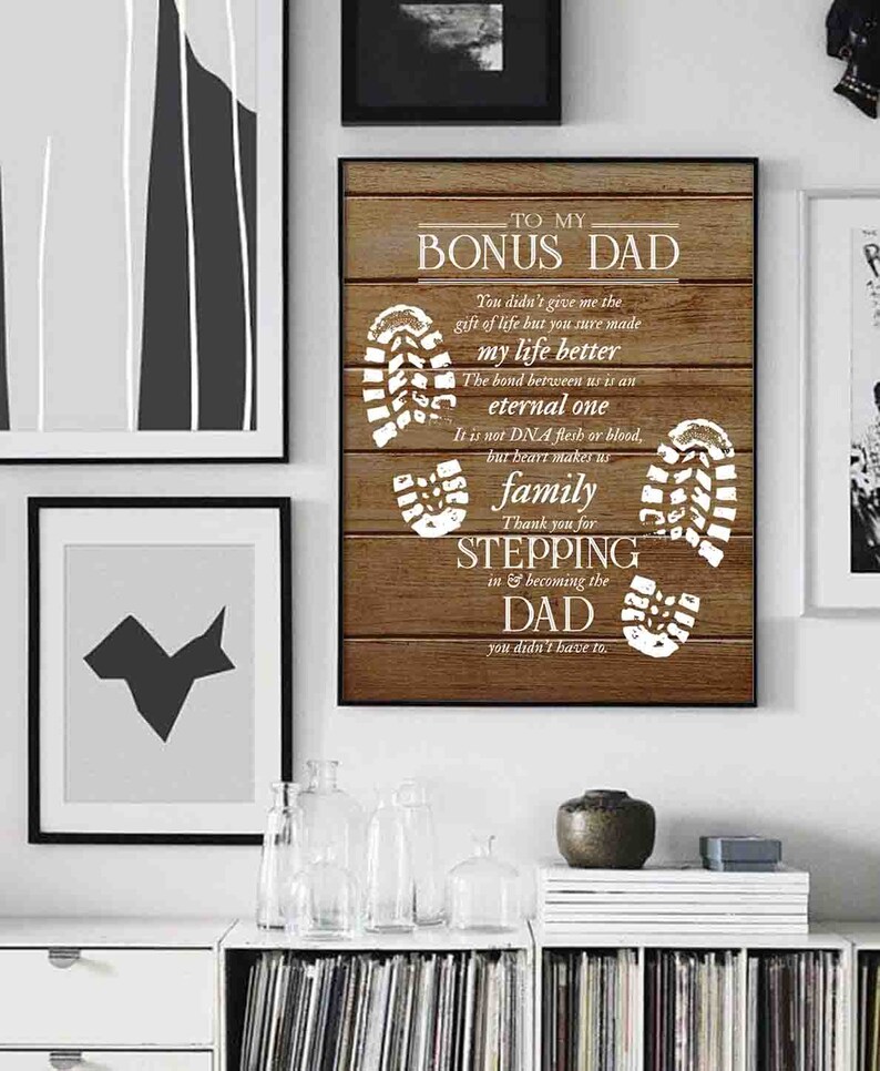 Puede incluir: Un letrero de madera con fondo blanco y textura de veta de madera marr&oacute;n. El letrero dice "To My Bonus Dad" e incluye un mensaje sobre el v&iacute;nculo entre un padrastro y un hijo. El letrero est&aacute; decorado con huellas blancas.