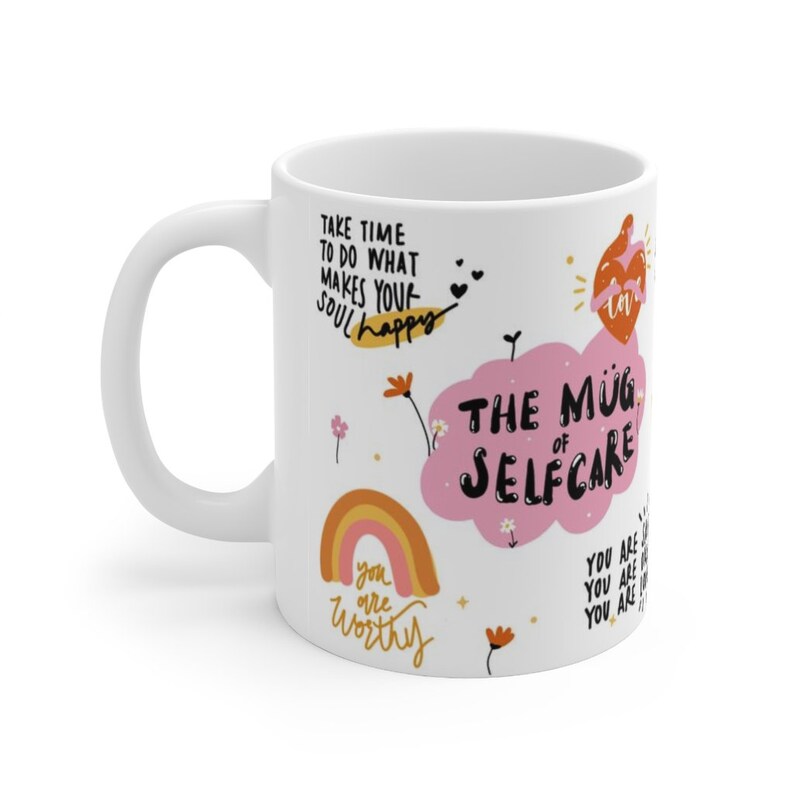 K&ouml;nnte beinhalten: Wei&szlig;e Keramiktasse mit einer rosa Wolke und dem Text "The Mug of Selfcare". Die Tasse zeigt au&szlig;erdem einen Regenbogen, Blumen und den Text "Take time to do what makes your soul happy" und "You are worthy".
