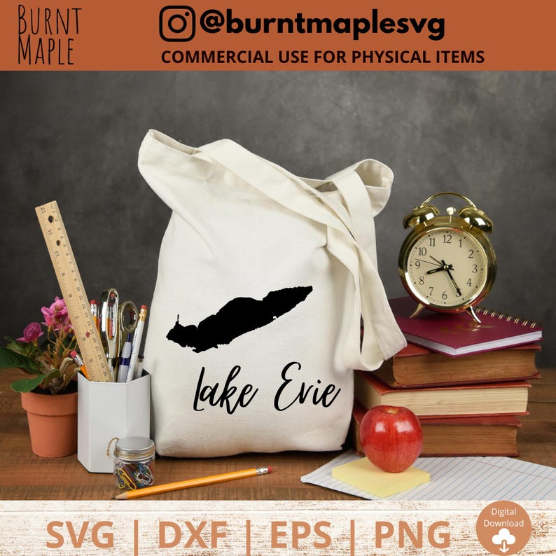 Puede incluir: Una bolsa de tela blanca con asas negras y el texto "Lake Evie" en escritura cursiva negra. La bolsa est&aacute; sobre una superficie de madera con una manzana roja, una pila de libros y un reloj despertador dorado.