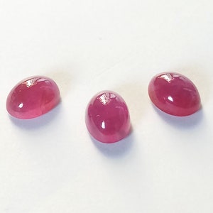 Peut inclure: Trois rubis de forme ovale, taillés en cabochon, d'une couleur rouge magenta profond avec une surface lisse et polie. Chaque pierre précieuse présente une qualité translucide avec des reflets lumineux sur les sommets arrondis.