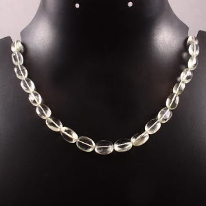 May include: A necklace composed of translucent, pale green, oval-shaped beads strung together. The beads have a smooth, polished finish and are arranged in a single strand, displaying subtle variations in clarity and light reflection throughout.