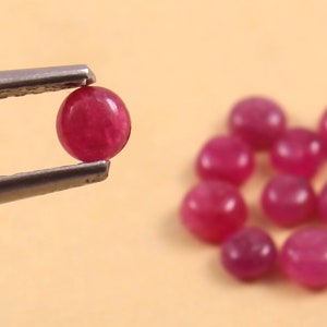 May include: A close-up of a ruby gemstone held in tweezers, with a scattering of similar ruby gemstones in the background.