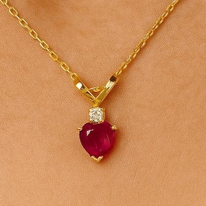 May include: A gold necklace with a heart-shaped, deep red pendant. The pendant is topped with a small, square, clear stone. The necklace chain is delicate and hangs against a skin-toned background.