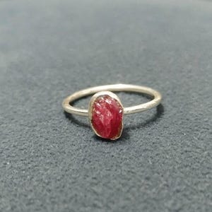 Puede incluir: Un anillo de plata con una gema de rubí en bruto, de forma ovalada. El rubí es de color rojo intenso y tiene una superficie texturizada. La banda del anillo es fina y de plata.