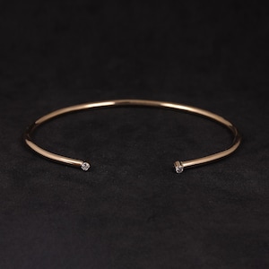 Può includere: Un bracciale aperto color oro con una piccola gemma rotonda e trasparente a ciascuna estremità. Il bracciale ha un design semplice ed elegante ed è posto su uno sfondo scuro.