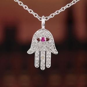 May include: A silver-toned Hamsa hand pendant necklace. The pendant is encrusted with small, clear stones and features a central pink gemstone. The necklace chain is also silver-toned.