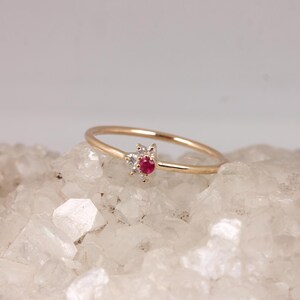 Può includere: Un delicato anello d'oro con una pietra rossa centrale circondata da pietre chiare più piccole. L'anello è posto su una superficie cristallina bianca. Il design è semplice ma elegante, con un focus sulle pietre preziose.