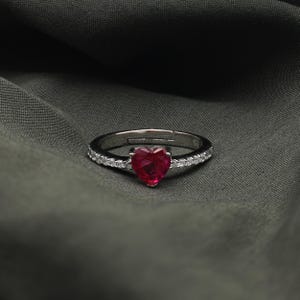 以下が含まれることがあります： 中央にハート型のルビーレッドの宝石が付いたシルバーリング。バンドには小さな透明な宝石が並んでいます。リングは濃い灰色の布の背景にセットされています。