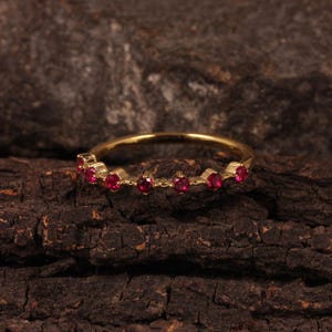 Op de afbeelding: Een gouden ring met een reeks kleine, ronde, rode edelstenen. De ring wordt getoond op een donkere, getextureerde houten ondergrond, die het contrast tussen goud en edelstenen benadrukt.