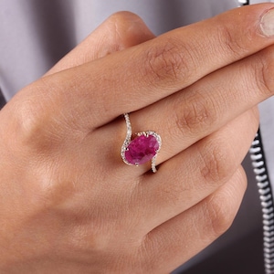 Könnte beinhalten: Ein Ring mit einem großen, oval geschliffenen, leuchtend rosa Edelstein, möglicherweise ein Rubin, der in einem zarten, wirbelnden Band aus weißem Metall, wahrscheinlich Silber oder Weißgold, gefasst ist. Das Band ist mit kleinen, funkelnden Akzentsteinen verziert.