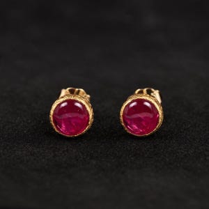 May include: A pair of gold stud earrings featuring round, vivid red gemstones. The earrings have a textured gold border, set against a dark backdrop, accentuating the jewellery's design.
