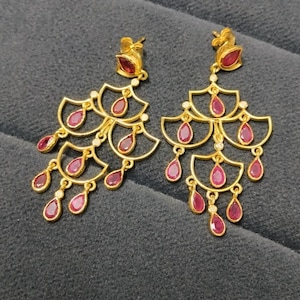 Pode incluir: Um par de brincos tipo candelabro em tom dourado com uma estrutura em camadas em forma de leque. Cada brinco é adornado com várias pedras vermelhas em forma de gota engastadas na estrutura metálica, criando um design simétrico em cascata.