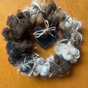 May include: A brown and white wool wreath with a black heart-shaped ornament in the center. The wreath is tied with twine and has a rustic look.