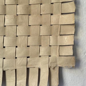 May include: Close-up of a woven paper artwork. The piece is constructed from beige paper strips, interlaced in a basket weave pattern. The edges of the paper are slightly frayed, adding texture. The artwork is displayed against a white background.