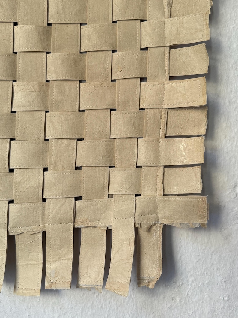 May include: Close-up of a woven paper artwork. The piece is constructed from beige paper strips, interlaced in a basket weave pattern. The edges of the paper are slightly frayed, adding texture. The artwork is displayed against a white background.