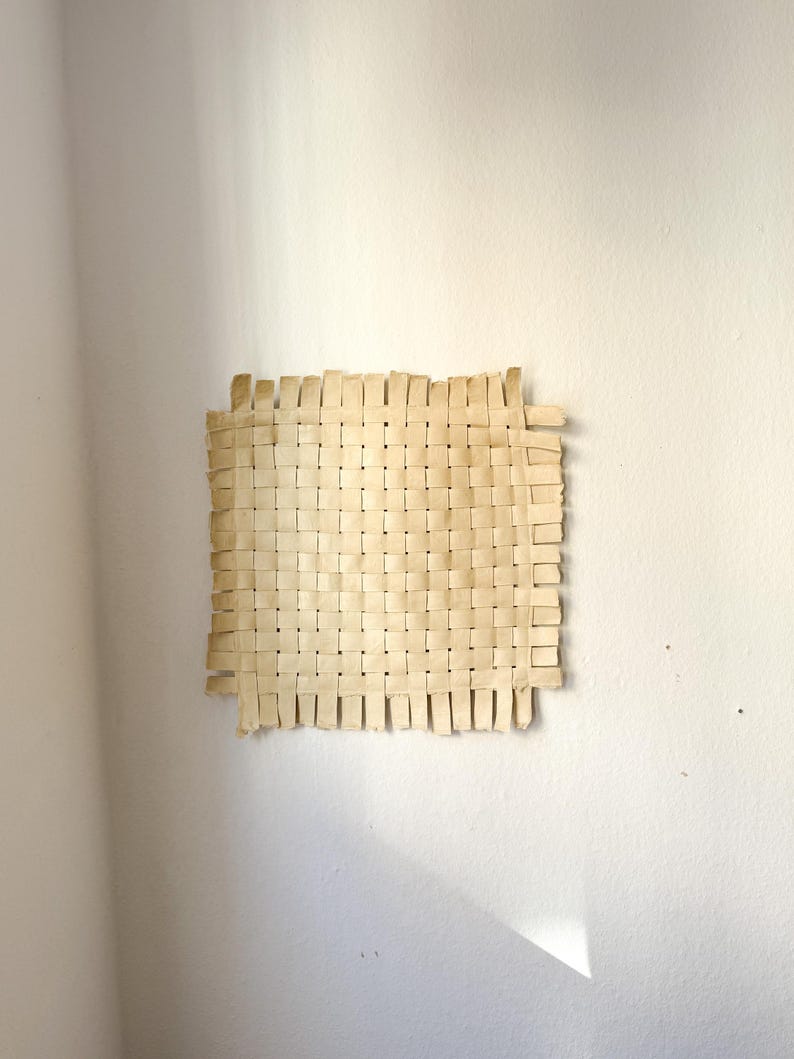 May include: A square, woven wall hanging made of light tan material. The weave pattern is visible, with vertical and horizontal strips interlaced to create a textured surface. The edges are slightly frayed, adding to the handmade aesthetic. It is mounted on a white wall.