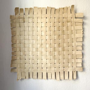 May include: A square, woven artwork made of light beige paper strips. The paper is woven in a basket-like pattern, with the edges of the paper strips visible. The artwork is mounted on a white wall.