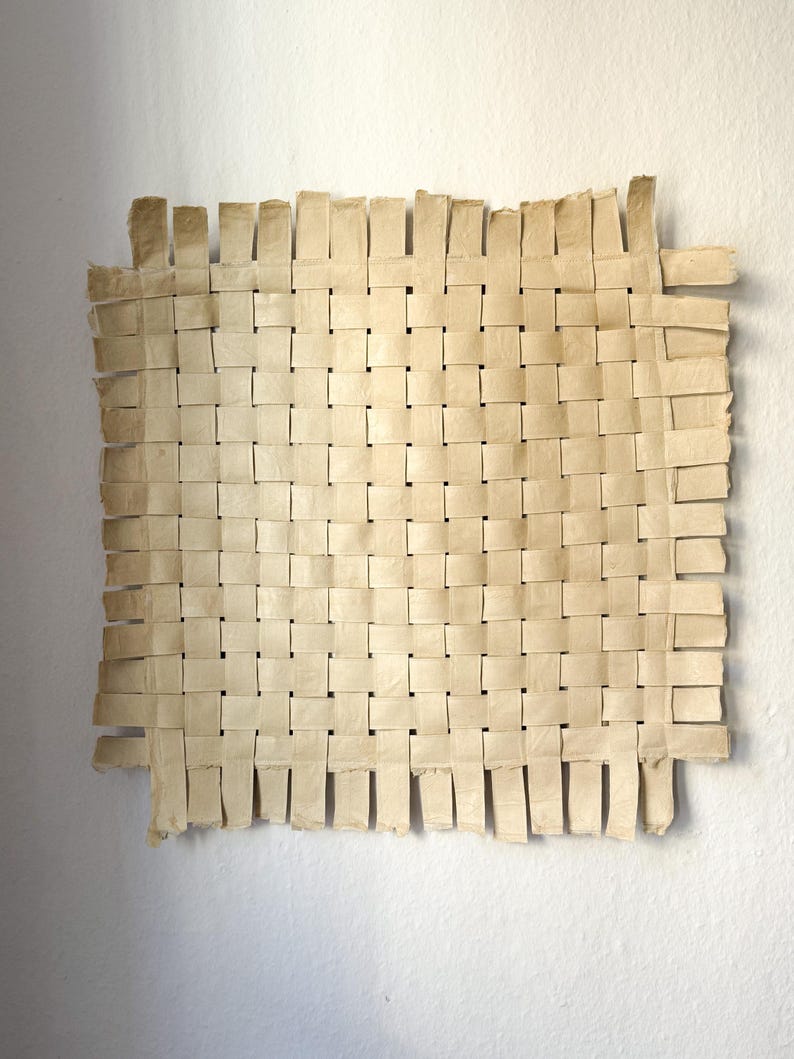 May include: A square, woven artwork made of light beige paper strips. The paper is woven in a basket-like pattern, with the edges of the paper strips visible. The artwork is mounted on a white wall.