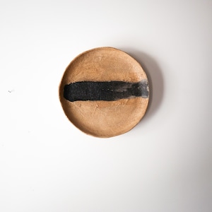 May include: A round, handmade ceramic plate with a textured, tan surface. A thick, horizontal band of black paint is brushed across the center. The plate is displayed against a white background.