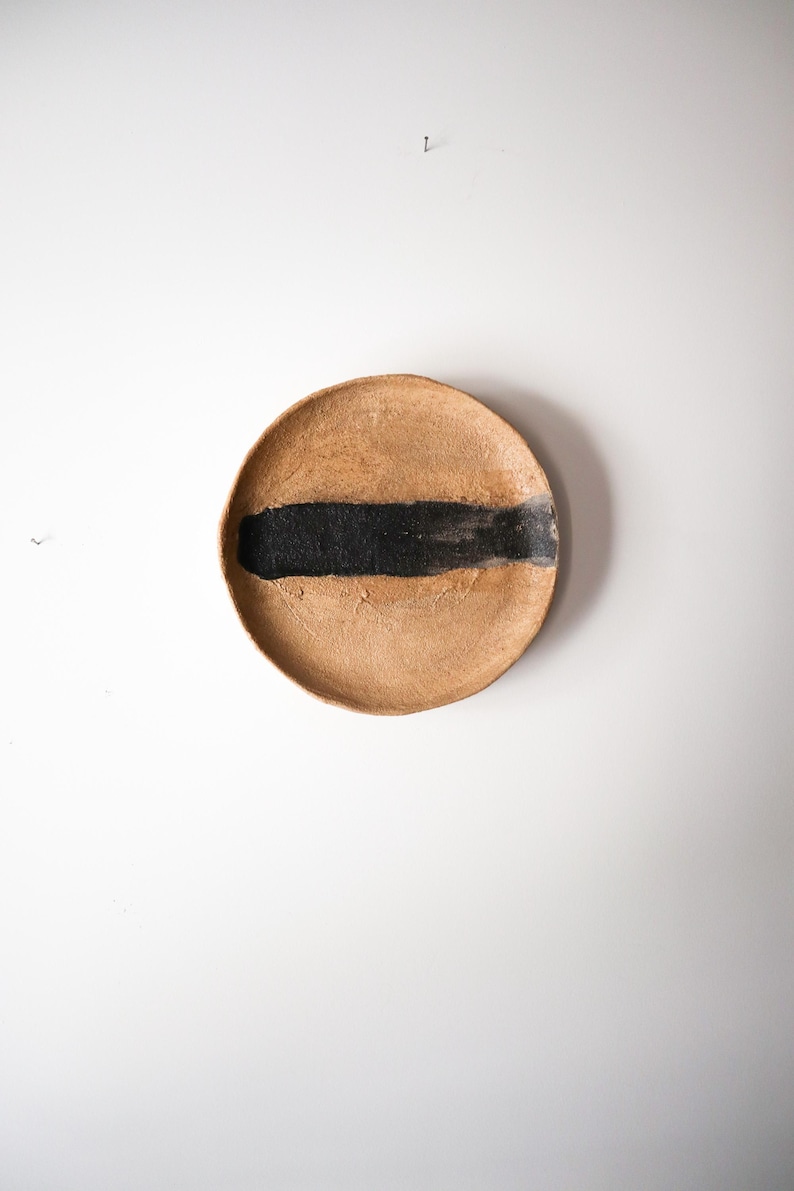 May include: A round, handmade ceramic plate with a textured, tan surface. A thick, horizontal band of black paint is brushed across the center. The plate is displayed against a white background.