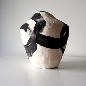 Peut inclure: Un vase en céramique beige avec des coups de pinceau noirs abstraits. Le vase a une forme arrondie et organique et est exposé sur une surface blanche. La peinture noire crée un contraste saisissant avec le fond neutre.