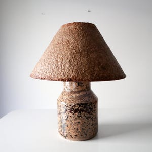 Pode incluir: Candeeiro de mesa de cerâmica castanha com abajur texturizado. O candeeiro tem uma base cilíndrica com um padrão salpicado em tons de castanho e bege. O abajur é cónico e tem uma superfície rugosa e texturizada.