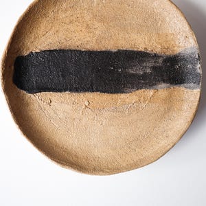 May include: A round, beige ceramic plate with a thick, horizontal black stripe across the center. The plate has a textured surface and a slightly raised edge. The design is minimalist and modern.