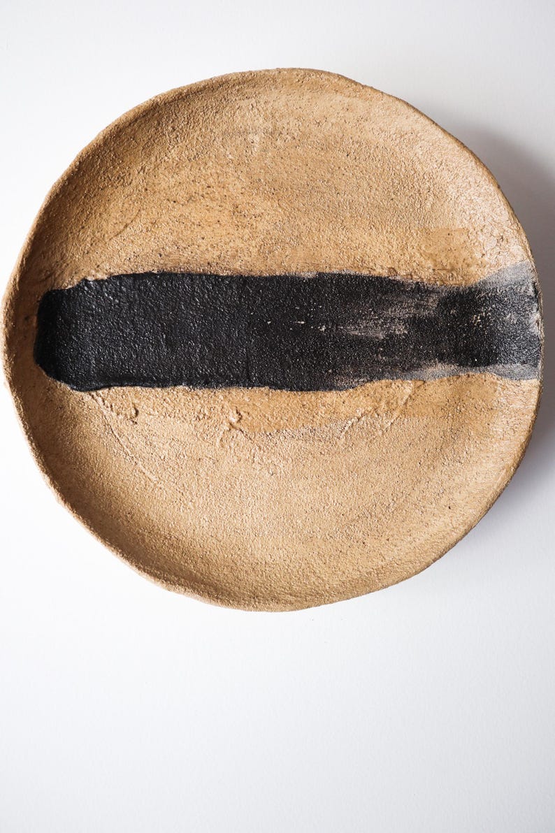 May include: A round, beige ceramic plate with a thick, horizontal black stripe across the center. The plate has a textured surface and a slightly raised edge. The design is minimalist and modern.