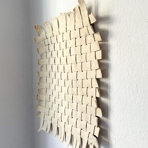May include: Off-white woven wall art with a rectangular shape and a curved edge. The woven pattern is made of rectangular strips, creating a textured surface. The artwork is mounted on a white wall, with shadows adding depth.