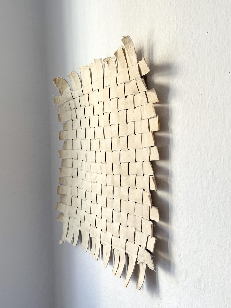 May include: Off-white woven wall art with a rectangular shape and a curved edge. The woven pattern is made of rectangular strips, creating a textured surface. The artwork is mounted on a white wall, with shadows adding depth.