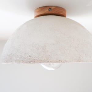 May include: A white, textured, dome-shaped ceiling light fixture with a wooden base. The light bulb is visible below the shade. The fixture is mounted on a white ceiling, providing ambient lighting.