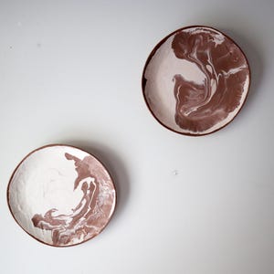 Peut inclure: Deux assiettes en céramique circulaires présentant un motif marbré avec des tourbillons de blanc et de brun terre cuite. Chaque assiette possède un bord lisse légèrement irrégulier et une finition brillante, révélant un mélange de couleurs.