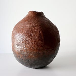 Peut inclure: Un vase en céramique marron fait à la main avec une surface texturée. Le vase a un corps arrondi et une petite ouverture en haut. Le bas du vase est d'une teinte plus foncée de marron.