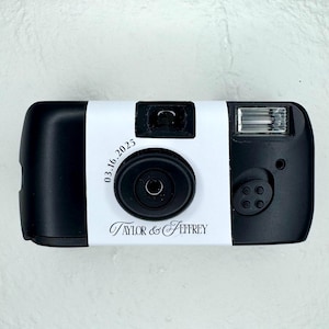 May include: A black and white disposable camera with a white band. The band features the date "03.16.2025" and the names "Taylor & Jeffrey" in elegant script. The camera has a flash and a lens.