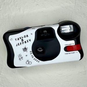 May include: A black and white disposable camera featuring the names "Taylor & Jeffrey" and the date "03.16.2025". The camera has a red flash button and wedding-themed illustrations, including a disco ball and champagne flutes.