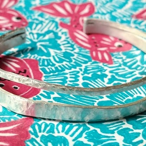 May include: Two silver hammered bangle bracelets stacked on top of each other. The bracelets are on a colourful patterned background.