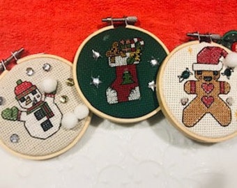 May include: Three handmade Christmas ornaments featuring a snowman, a stocking, and a gingerbread man. The ornaments are stitched on white, green, and brown fabric and are framed in wooden hoops.