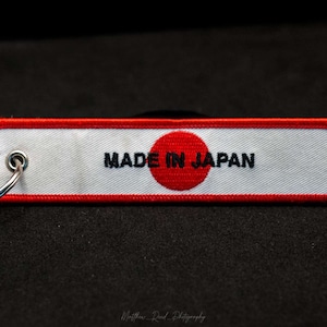 May include: A white keychain with red trim and a red circle with a white outline. The keychain says "MADE IN JAPAN" in black text.