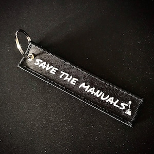 May include: Black fabric keychain with the text "SAVE THE MANUALS" in white, along with a small graphic. The keychain has a metal ring for attaching keys. The design is simple and bold.