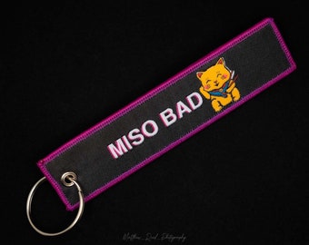 May include: A black and pink key chain with a white embroidered text "MISO BAD" and a yellow cartoon cat with a knife.