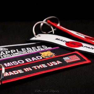 May include: Embroidered keychains with various designs. One keychain reads "MADE IN THE USA" with an American flag. Another says "MADE IN JAPAN" with a red circle. Other keychains have text and a cartoon cat.