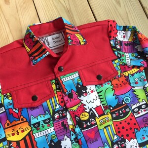 Peut inclure: Une veste en jean rouge avec une doublure à imprimé chat coloré. La veste a une fermeture à boutons et deux poches. L'étiquette sur le col indique "Louise Beland".