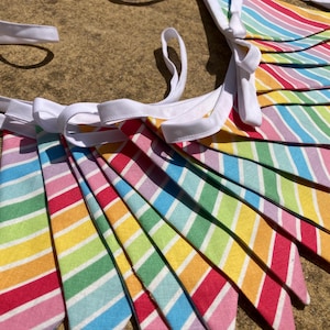 May include: A rainbow striped bunting banner with white ribbon ties. The pennants feature stripes of red, pink, orange, yellow, green, blue, and purple. The fabric banner is ideal for party decorations and measures approximately 150 cm in length.