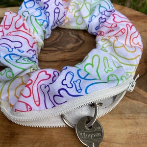 Könnte beinhalten: Ein weißer Haargummi mit einem Regenbogenmuster aus abstrakten Formen und einem Reißverschluss. Ein silberner Schlüssel mit dem Text "Yale", "Timpson" ist am Reißverschluss befestigt.
