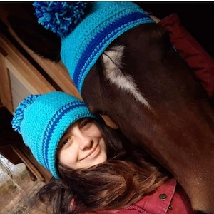 May include: A woman and a horse wearing matching turquoise and blue crocheted beanies with pom-poms. The woman is smiling, and the horse has a dark brown coat. The beanies have a striped design.