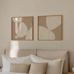 May include: Two framed abstract art prints with a beige background and white shapes. The prints are hanging on a wall above a beige headboard with white and beige pillows.