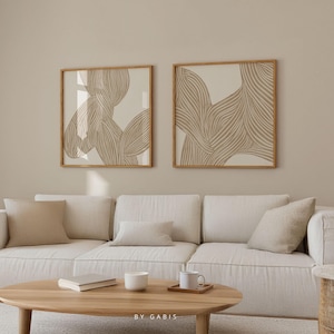 Puede incluir: Dos impresiones de arte abstracto enmarcadas con líneas beige y marrones sobre un fondo blanco. Las impresiones están colgadas en una pared beige sobre un sofá blanco con almohadas beige.