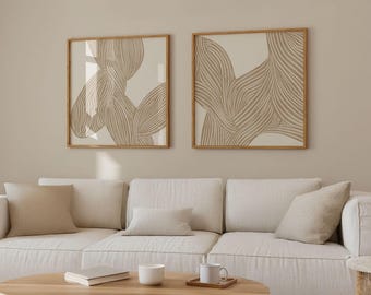 May include: Two framed abstract art prints with beige and brown lines on a white background. The prints are hanging on a beige wall above a white couch with beige pillows.