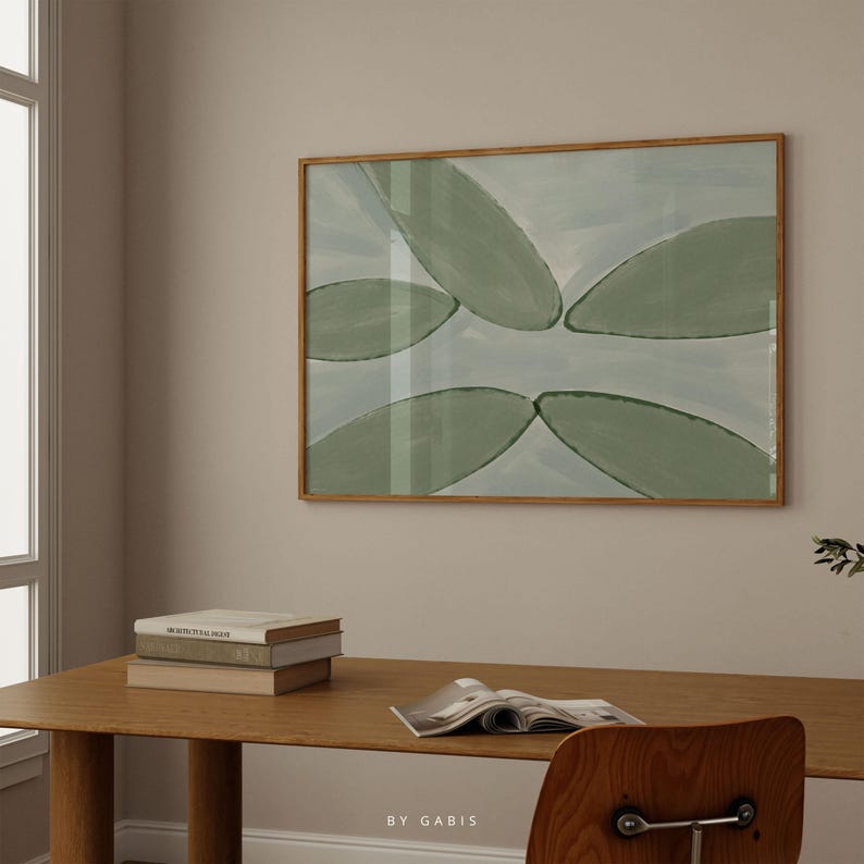 May include: A framed abstract painting with a light blue background and green leaf shapes. The painting is hanging on a light beige wall above a wooden table with books and a magazine.