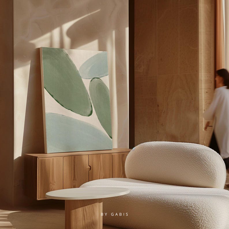 May include: Abstract painting featuring green and blue shapes on a white background. The painting is displayed on a wall in a room with a wooden cabinet and a white sofa.